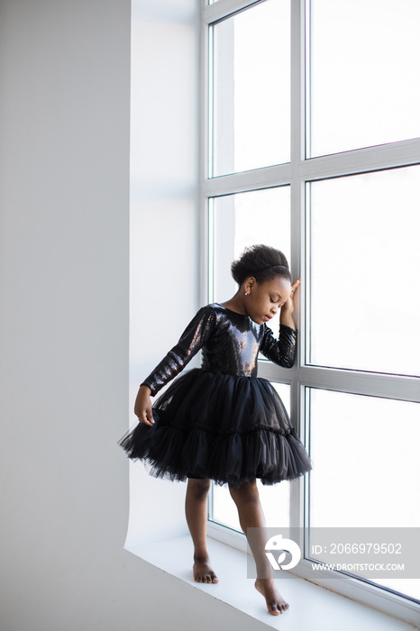 Tender little female kid in elegant black dress posing barefoot at studio with white background. Pretty african ballerina standing near window and looking down.