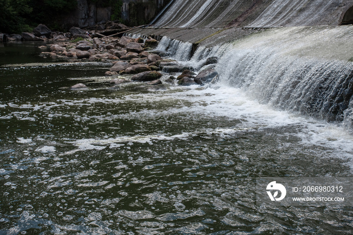 Picturesque textured waterfall on the river. There are big rocks along the river. Image of falling water on a dam, as a background for your design or illustrations.