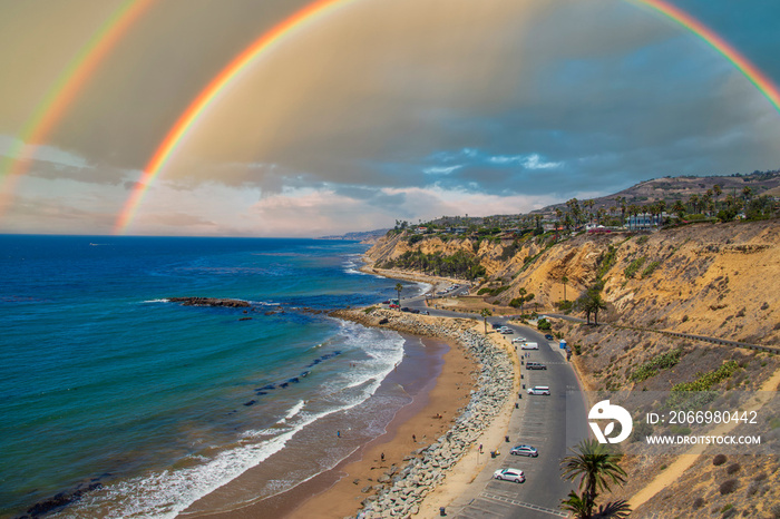 a gorgeous summer landscape along the coastline with blue ocean water and cliffs covered with lush green palm trees, grass and plants with blue sky and a rainbow and waves rolling into the beach