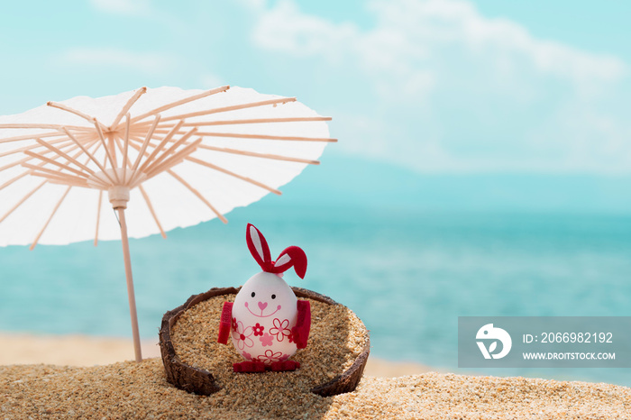 Creative concept for celebrating Easter on beach. Easter egg on coconut shell on golden sandy beach under sun umbrella and blurred blue sea and sky in background. Greeting card, copy space