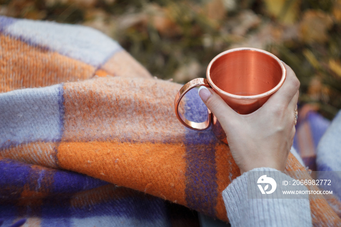 Close-up female hand holding a shiny copper mug with tea on the background of a bright woolen plaid, cozy autumn concept, selective focus