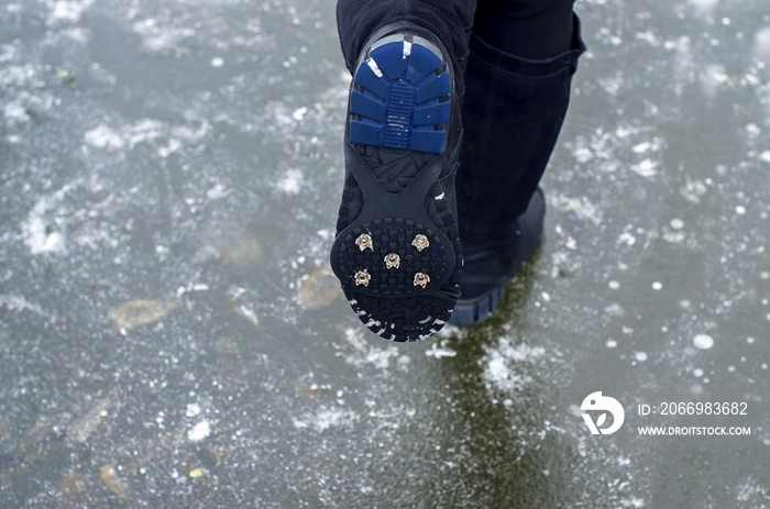 Female legs in black boots with Ice Grippers Cleats for Shoes and Boots on slippery road with frozen puddle covered with ice. Concept of injury risk in winter and danger