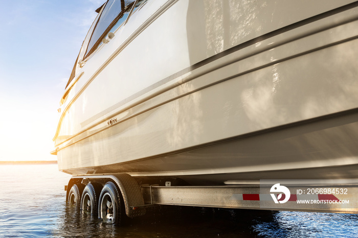 Big luxury cabin motorboat cruiser yacht launching at trailer ramp on river or lake. Warm morning sunrise sunshine reflection in calm water surface. Luxury rich fishing leisure recreation lifestyle