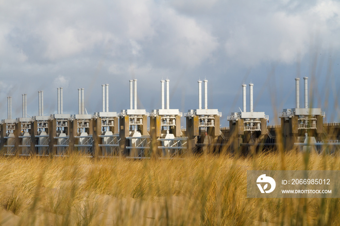 View across the sea to the Eastern Scheldt Storm Surge Barrier in Zeeland with a stormy sky, dramatic sunlight with sand dunes, grass and beach in foreground. Oosterscheldekering during winter season