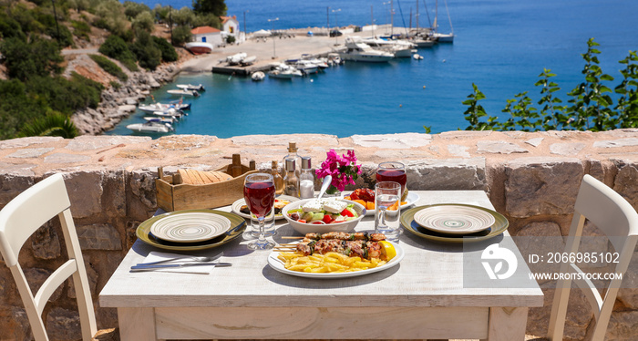 A table served for two with chicken souvlaki and french fries, greek salad, snacks and drinks on the summer terrace of the hotel room by the seascape, beautiful summer greek holidays concept.