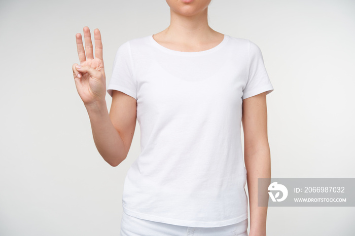 Cropped shot of young female’s hand with nude manicure showing three fingers while meaning letter w, standing over white background in casual clothes