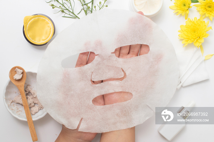 woman hand holding white sheet mask with clay mask wooden spoon yellow flowers and serum skincare cream products on white background