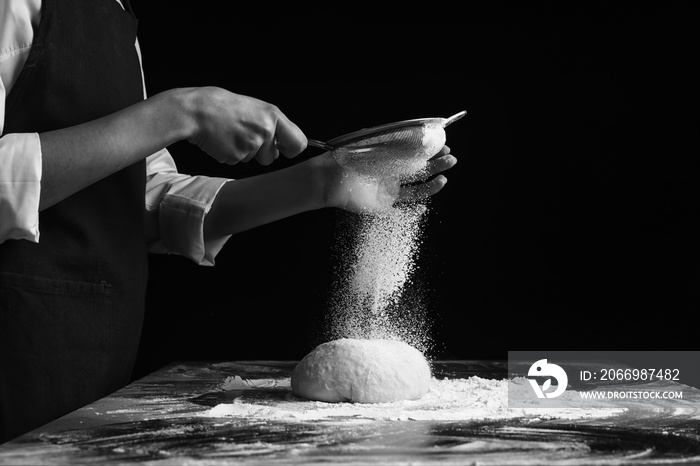 Woman sieving flour on dough on dark background