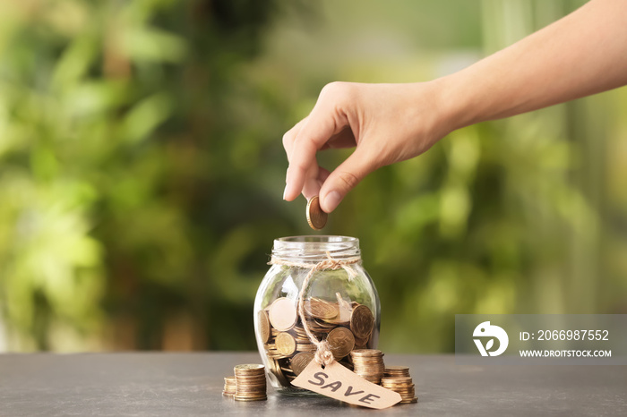 Woman putting coin into glass jar against blurred background