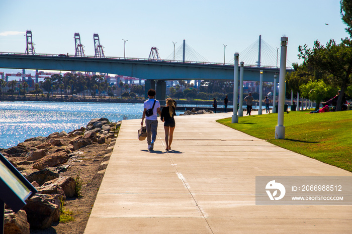 a couple walking along a smooth bike path at the park along the blue ocean water surrounded by lush green trees and grass with blue sky and a bridge at ShoreLine Aquatic Park in Long Beach California