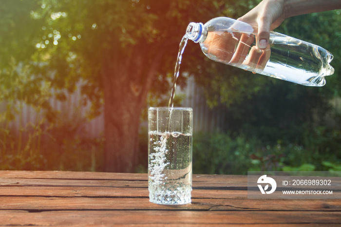 Female hand pours water from a plastic bottle into a glass on a wooden table in a spring garden