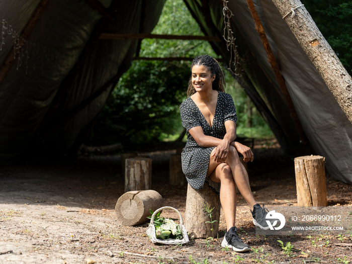 Smiling woman resting on tree stump in front of hut in forest