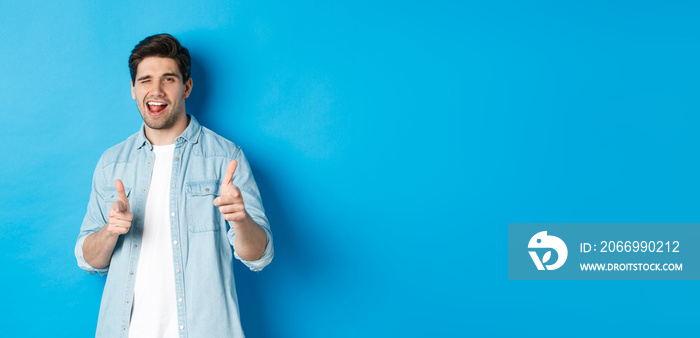 Confident adult man winking, pointing finger at you and winking, congratulating or praising person, standing against blue background