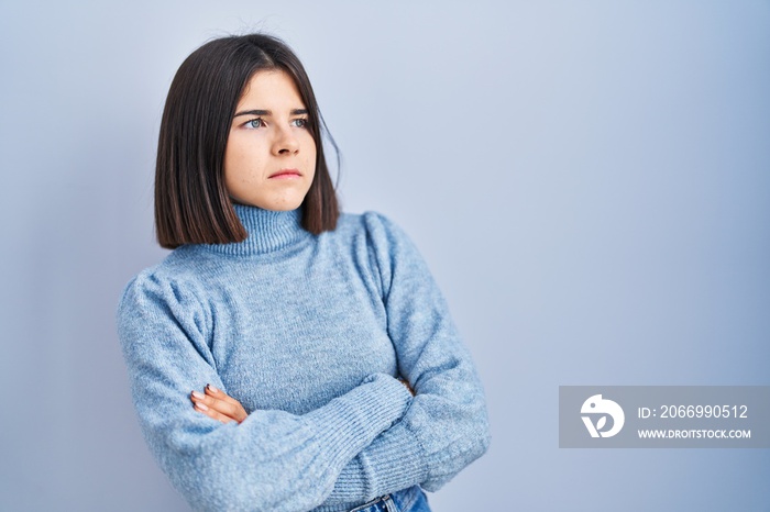 Young hispanic woman standing over blue background looking to the side with arms crossed convinced and confident