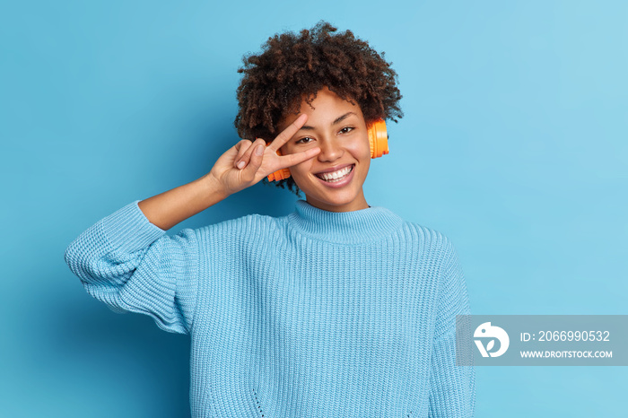 Cheerful dark skinned curly woman listens favorite music in headphones has toothy smile makes victory gesture wears casual sweater isolated over blue background. People and lifestyle concept