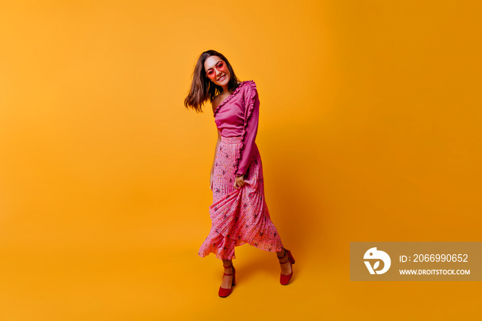 �ntented, glamorous young woman in fashionable pink clothes demonstrates her outfit in all its glory. Brunette in colored glasses smiles sweetly, looking at camera on orange background