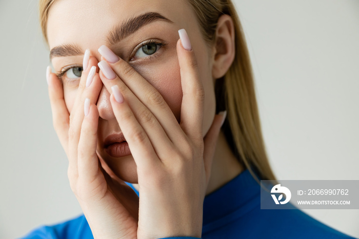 young blonde woman in blue turtleneck obscuring face with hands and looking at camera isolated on grey.