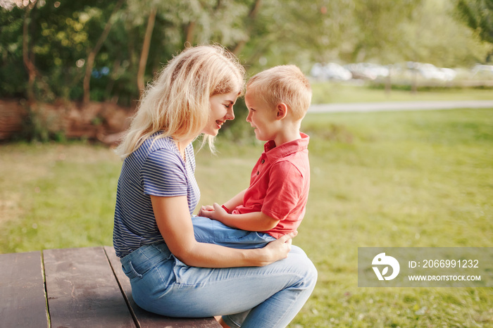 Young Caucasian mother and boy toddler son sitting together face to face. Family mom and child talking communicating outdoor on a summer day. Happy authentic family childhood lifestyle.