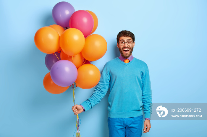 Positive attractive man with happy look, wears round glasses, blue outfit, celebrates festive event, holds colorful balloons, poses indoor. Delighted guy comes on stag party, has fun with friends