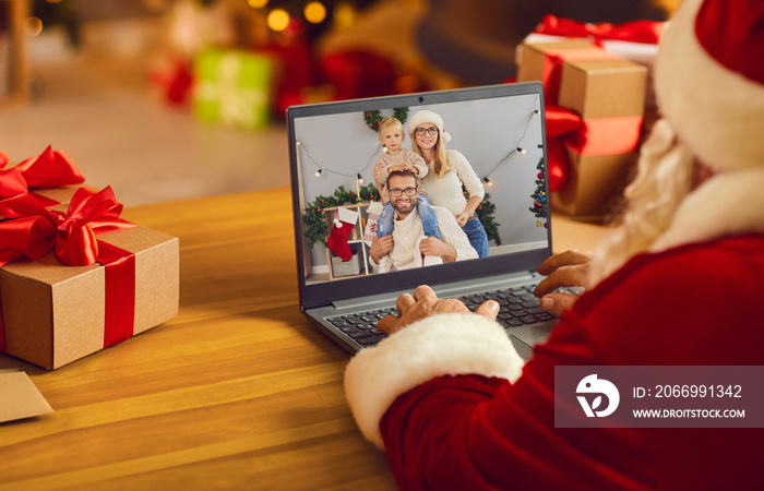 Santa Claus in his workshop video calling young family using modern laptop computer