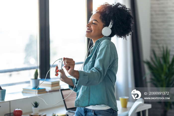 Motivated young afro woman listening to music with headphone while dancing in living room at home.