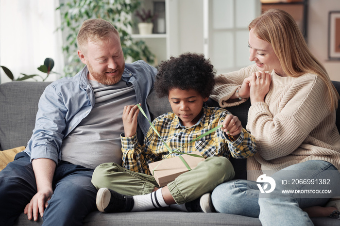 African little boy opening gift box giving by his foster parents while they sitting on sofa in the room