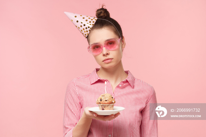 Unhappy birthday girl holding plate with holiday cake and looking at it with sad face, tired of party, isolated on pink background