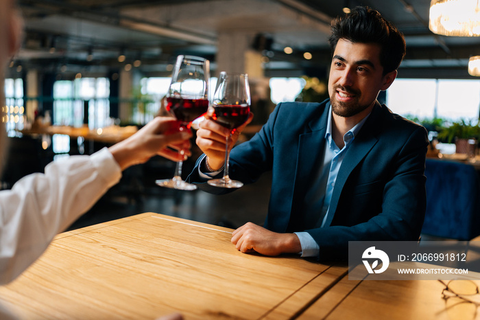 View from back of unrecognizable female clinking glasses of red wine with smiling elegant man in suit sitting at table in fancy restaurant at evening. Happy young couple enjoying nice romantic dinner.
