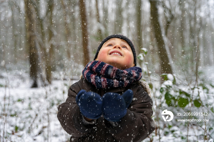 Close up portrait of a little boy playing with snowflakes in a park in winter. Happy child enjoys the first snow in a forest.