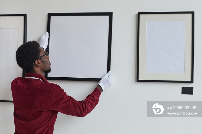 Back view at African-American man hanging frames on wall while planning art gallery or exhibition, copy space