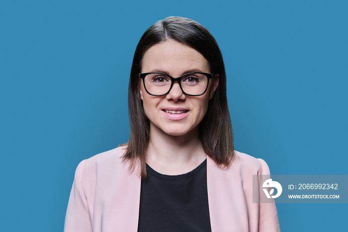 Headshot portrait of young woman in glasses looking at camera on blue background