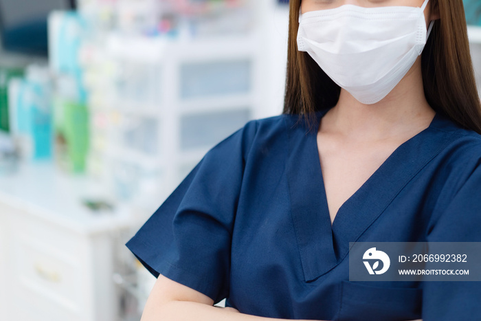 Cropped view and focus of young long-haired Asian female nurse or doctor in a blue uniform wearing a white medical face mask to protect Covid-19 standing with arms folded in a blurred hospital room.