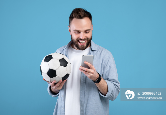 Happy young guy with soccer ball using smartphone, winning sports bet, rooting for his favorite team on blue background