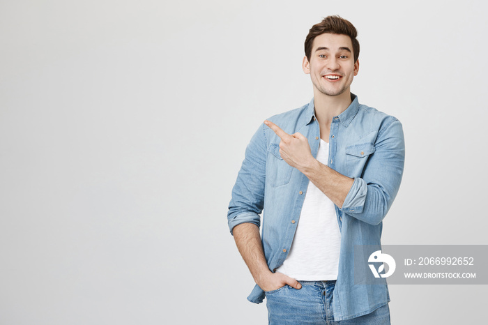 Studio portrait of attractive european man in denim glothes, pointing at upper left corner with index finger, expressing excitement, happiness and surprise, over gray background.