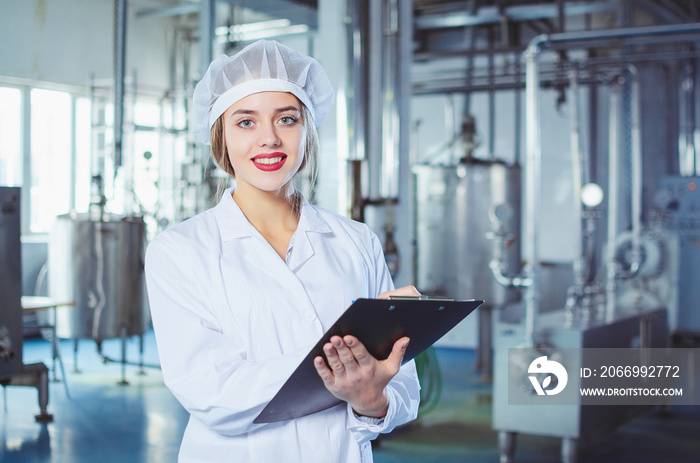 A young beautiful girl in white overalls makes notes in a tablet on the background of equipment of a food processing plant. Quality control in production