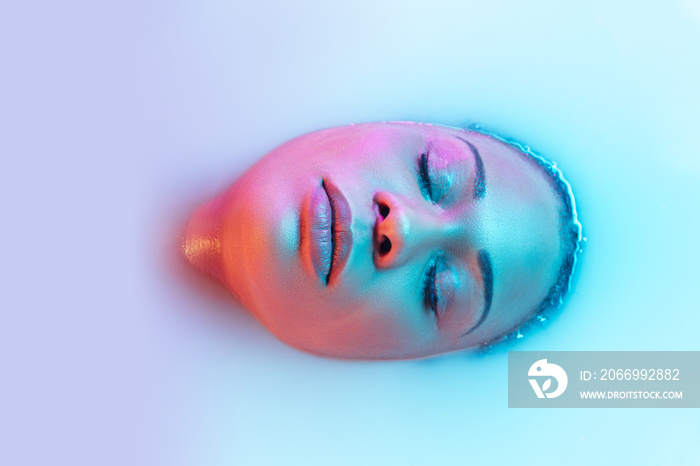 Eyes closed. Beautiful female face in the milk bath with soft glowing in blue-pink neon light. Copyspace for advertising. Modern neoned colors, foam. Beauty, fashion, style, skincare concept