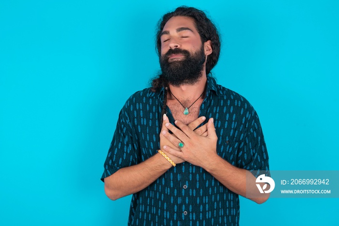 young bearded man wearing blue shirt over blue stud closes eyes and keeps hands on chest near heart, expresses sincere emotions, being kind hearted and honest. Body language and real feelings concept.