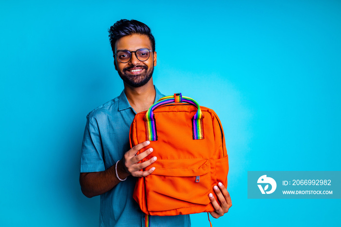 handsome young bearbed indian man with eye glasses in blue cotton t-shirt with orange rainbow backpack in studio background