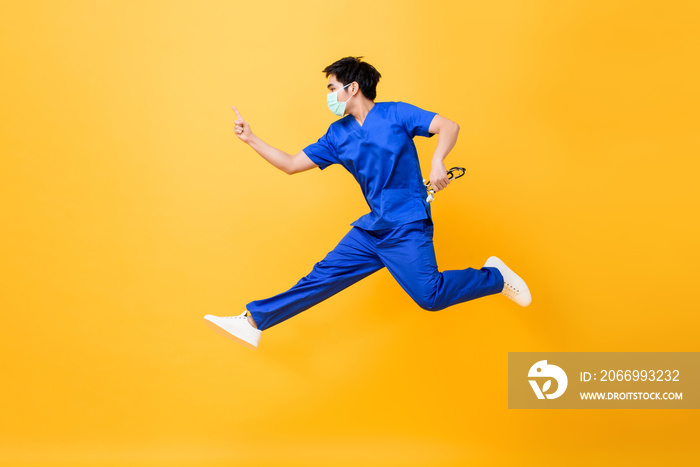 Young Asian male doctor wearing medical mask jumping and pointing hand up isolated in yellow studio background