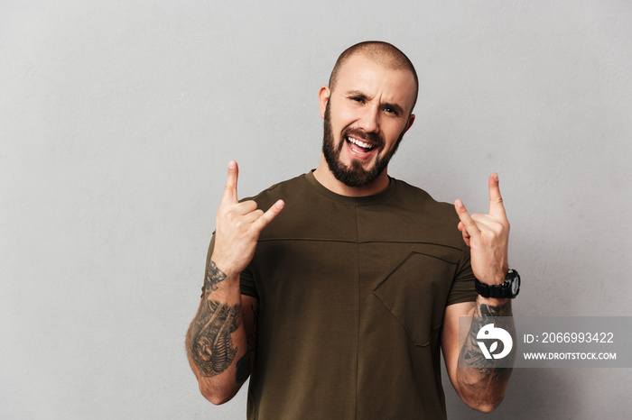 Image of cheerful hairless guy with beard and mustache smiling and showing rock sign, isolated over gray background