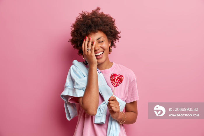 Positive African American teenage girl makes face palm, has fun, dressed in casual clothes, holds heart shaped lollipop, enjoys spare time, poses against pink background. Confectionery concept