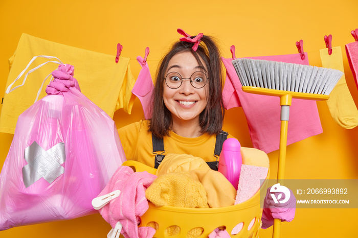 Positive Asian woman holds broom for sweeping floor poses with cleaning supplies polythene bag full of detergents does laundry at home busy doing housework and domestic duties. Housecleaning