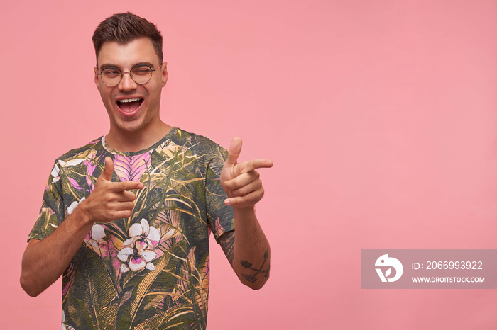 Indoor shot of enthusiastic attractive guy with glasses, indicating with fingers aside, giving wink, isolated on pink background