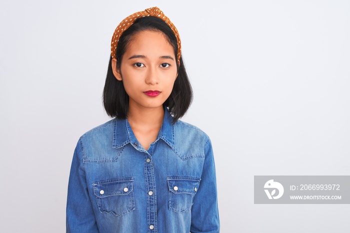 Young beautiful chinese woman wearing denim shirt standing over isolated white background with serious expression on face. Simple and natural looking at the camera.