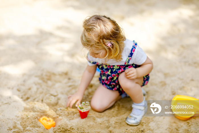 Cute toddler girl playing in sand on outdoor playground. Beautiful baby in red trousers having fun on sunny warm summer day. Child with colorful sand toys. Healthy active baby outdoors plays games