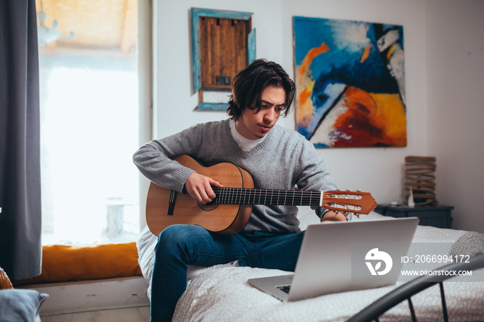 teenager playing guitar in his room at home, using laptop computer for online lessons