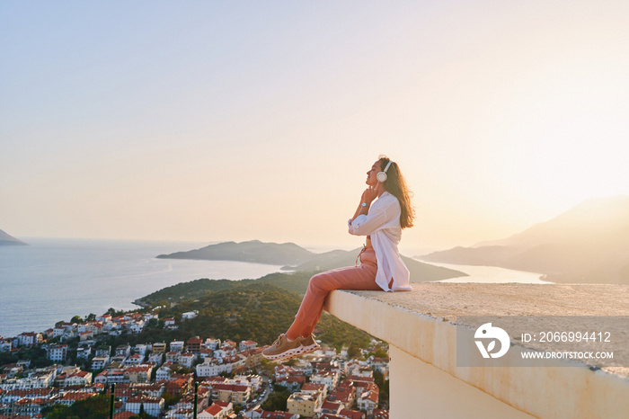 Inspired happy female sitting on top and enjoying of listening calm music with beautiful view at sunset time. Relief stress and healthy minds