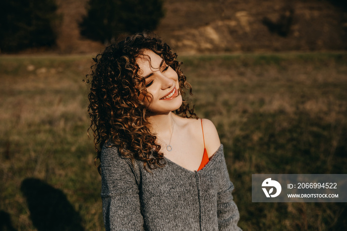 Portrait of a young woman with curly hair, smiling, outdoors, in sun light.