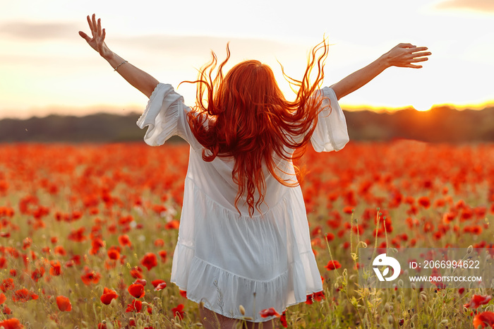 Happy redhead smiling woman in white dress on field of poppies at summer sunset