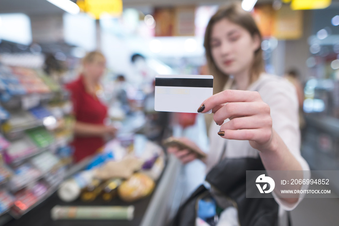 Woman is at the supermarket’s cash desk and shows a card in the camera. Payment by credit card at a supermarket. Card in the hands of a woman close-up on the background of the supermarket cash desk